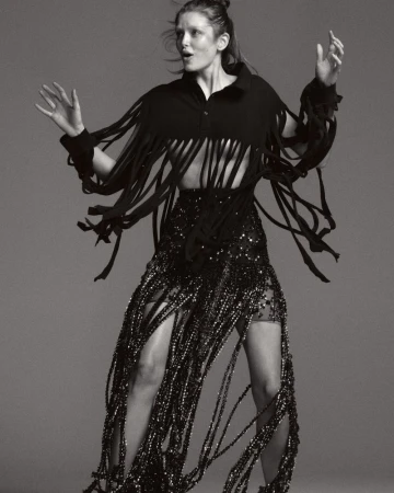 Adult woman in a black fringed top and skirt with sequins stands in a photo studio. Arms raised with a neutral facial expression.