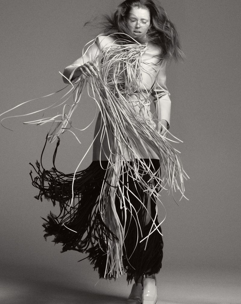 An androgynous fashion model in motion wears a white jacket and a skirt of white and black tassels against a solid gray backdrop.