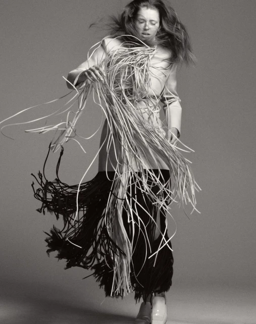 An androgynous fashion model in motion wears a white jacket and a skirt of white and black tassels against a solid gray backdrop.
