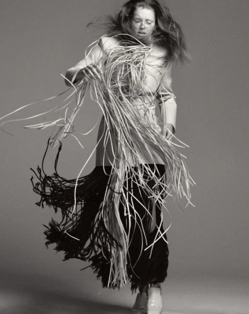 An androgynous fashion model in motion wears a white jacket and a skirt of white and black tassels against a solid gray backdrop.