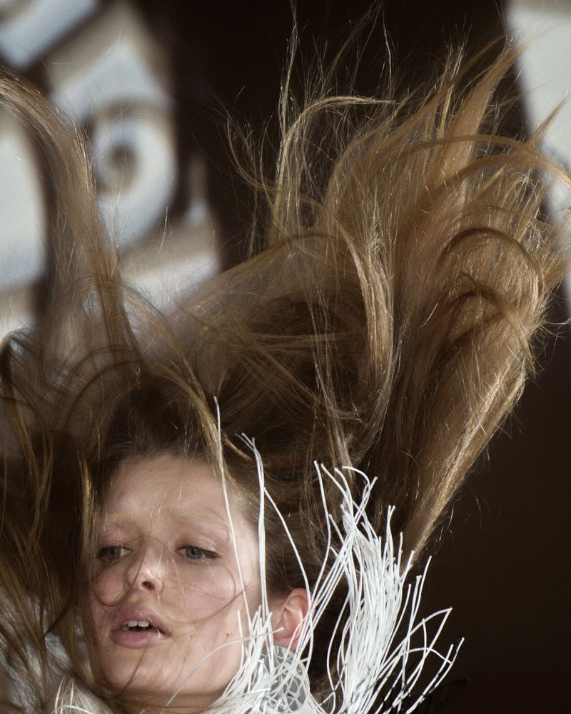 Blond-haired woman looking down with hair falling down around her face; she wears an outfit with string detail around her neck.