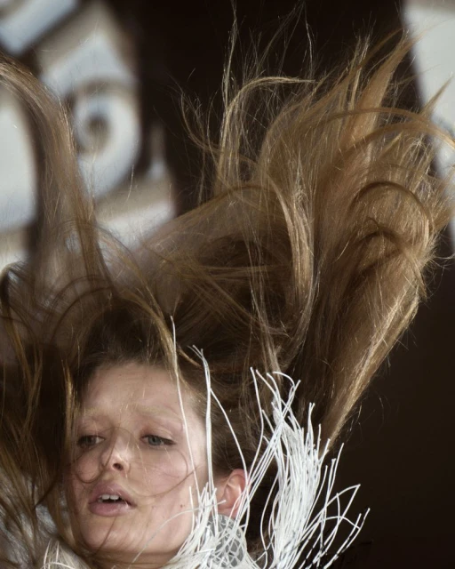 Blond-haired woman looking down with hair falling down around her face; she wears an outfit with string detail around her neck.