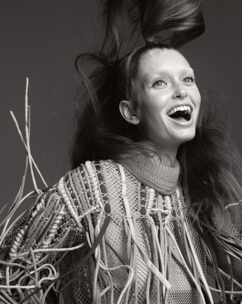 Portrait of a smiling young woman with her long brown hair flying upwards, wearing a mesh top with fringe, against a grey backdrop.
