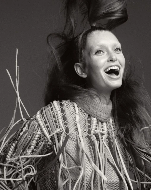 Portrait of a smiling young woman with her long brown hair flying upwards, wearing a mesh top with fringe, against a grey backdrop.