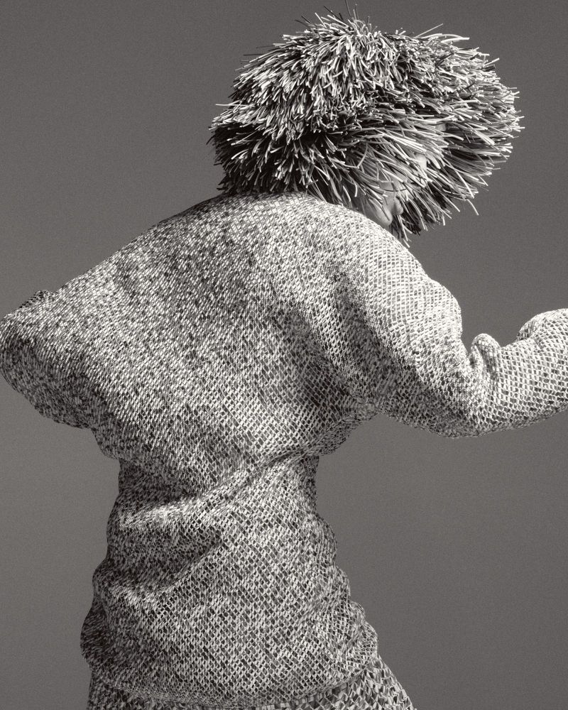 Monochrome fashion shot of an adult wearing a textured dress and an unusual headdress, posing with their back to the camera in a studio.