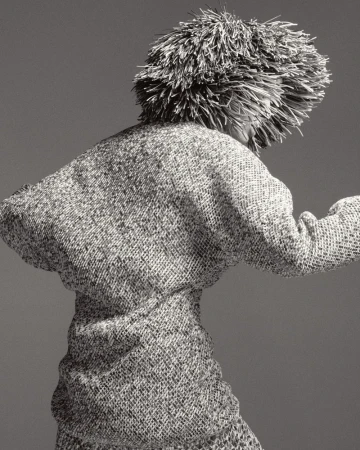 Monochrome fashion shot of an adult wearing a textured dress and an unusual headdress, posing with their back to the camera in a studio.