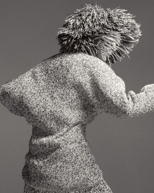 Model wearing a textured fringed hat and patterned knit garment against a solid grey background