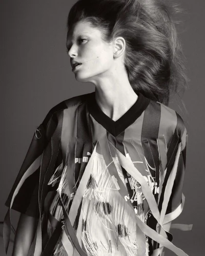 Black and white portrait of a female model wearing a fringed sports jersey with hair blowing against a grey background.