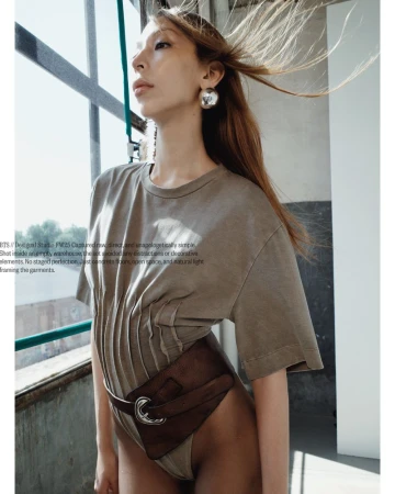 Indoor shot of a slim adult female model in a neutral bodysuit with brown belt bag, looking at window light. Hair is long and brown.