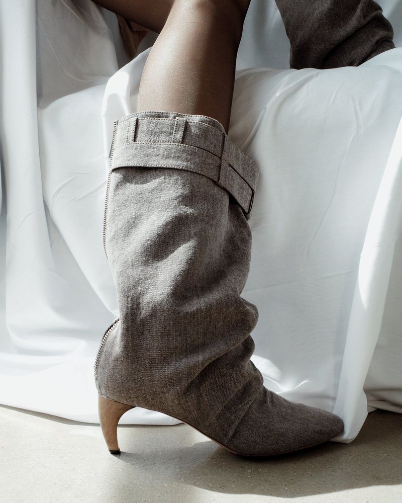 Grey knee-high slouch boot with buckle detail. The shoe has a curved heel and is paired with a white, draped fabric background.