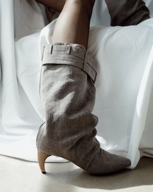 Grey knee-high slouch boot with buckle detail. The shoe has a curved heel and is paired with a white, draped fabric background.