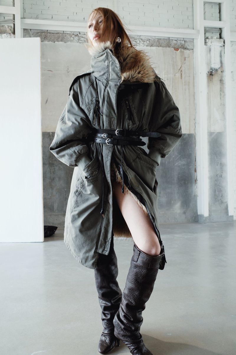 A young adult woman with fair skin poses in a fur-trimmed olive coat cinched with a wide black belt and high leather boots in a studio.