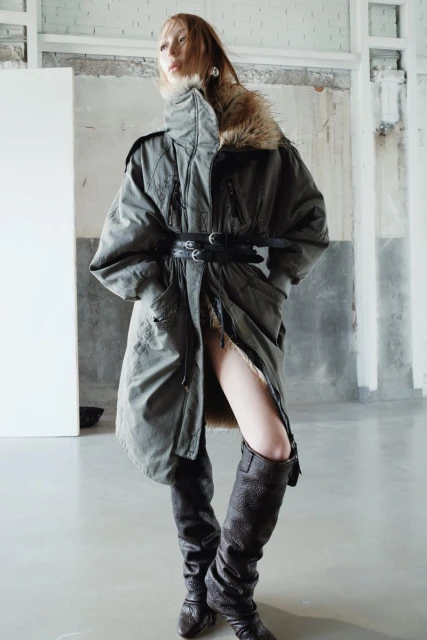 A young adult woman with fair skin poses in a fur-trimmed olive coat cinched with a wide black belt and high leather boots in a studio.