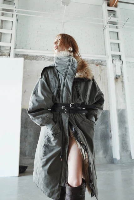 A stylish woman in a green parka coat with a fur collar and a black belt, posing in a bright, minimalist interior.
