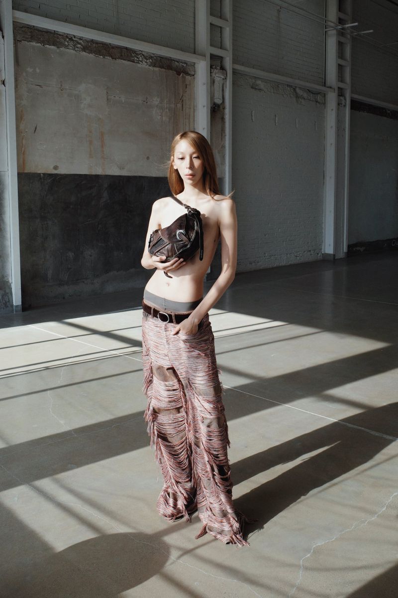 Adult female poses in a bright industrial interior with a dark-brown crossbody bag, and fringed trousers in bright, soft window lighting.
