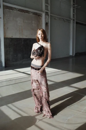 Adult female poses in a bright industrial interior with a dark-brown crossbody bag, and fringed trousers in bright, soft window lighting.