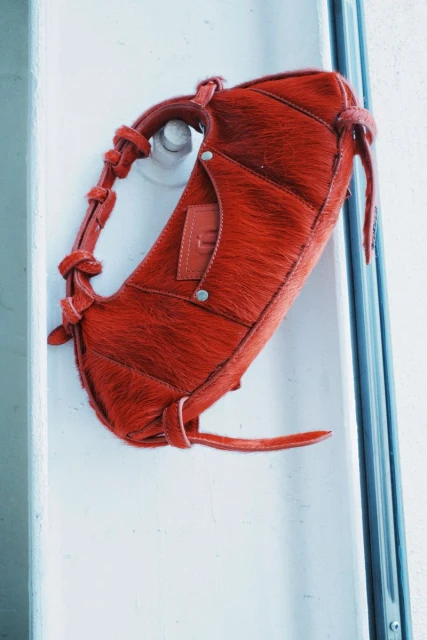 Close-up of a small, red, furry handbag with leather detailing hanging on a white door with a silver handle, fashion accessory.