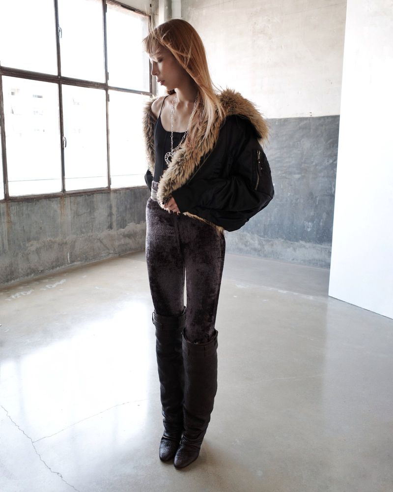A young woman poses in an indoor space wearing a black jacket with fur trim, velvet pants, and high boots near a bright window.