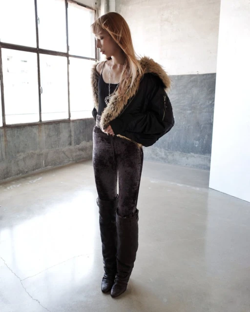 A young woman poses in an indoor space wearing a black jacket with fur trim, velvet pants, and high boots near a bright window.