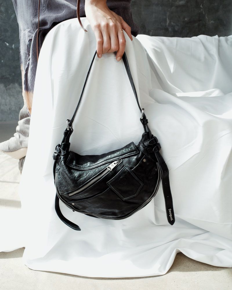 Black leather shoulder bag held by a person in a casual outfit against a wrinkled white backdrop. Fashion shot, neutral background.