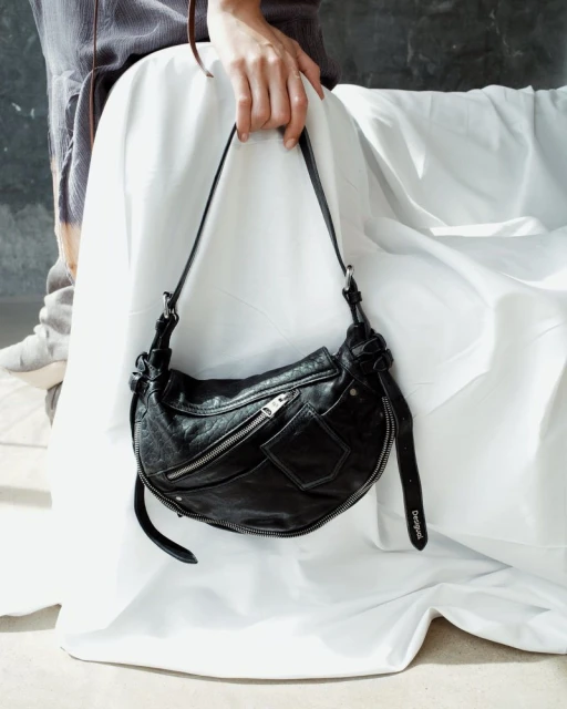 Black leather shoulder bag held by a person in a casual outfit against a wrinkled white backdrop. Fashion shot, neutral background.