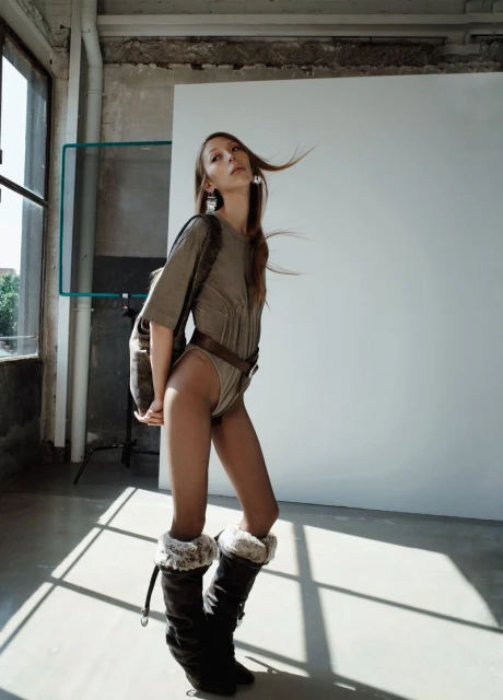 Full-length shot of a slender young woman posing indoors wearing a bodysuit, belt, backpack, and boots against a white backdrop.