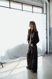 Model standing in profile against a bright white studio backdrop wearing a dark patterned jacket and loose trousers.