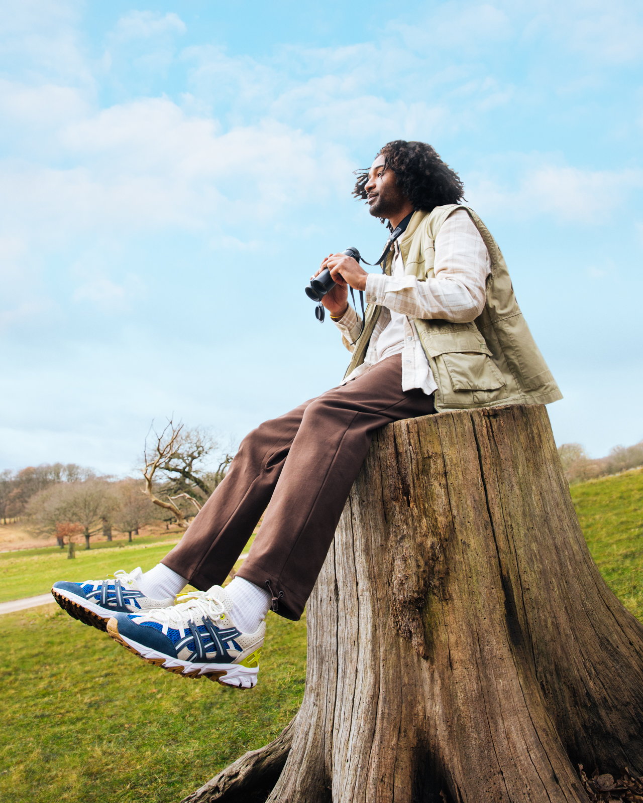 Adult male sitting on a tree stump with binoculars in hand in nature with a blue sky in the background and wearing trainers.