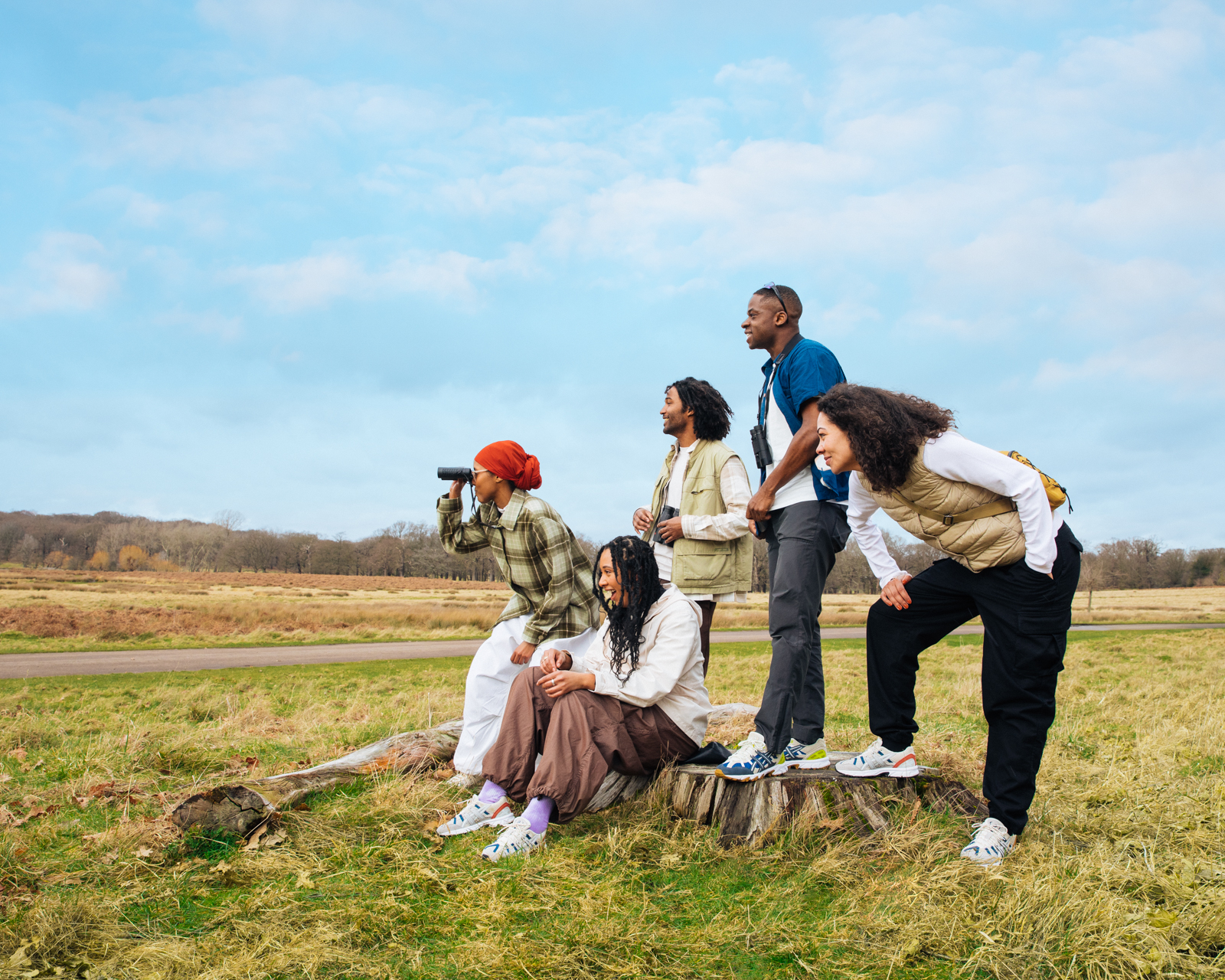 A diverse group of five adults casually poses on a tree stump in an open grassy field under a bright blue sky with binoculars.