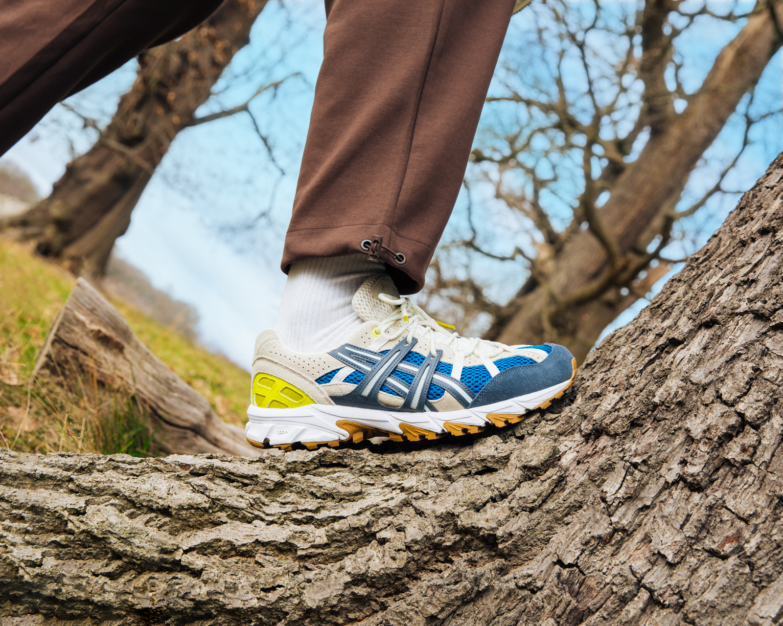 Close-up of a person's legs and feet stepping onto a tree trunk with Asics sneakers outdoors in a forest setting with a blue sky.