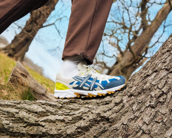 Close-up of a person's legs and feet stepping onto a tree trunk with Asics sneakers outdoors in a forest setting with a blue sky.