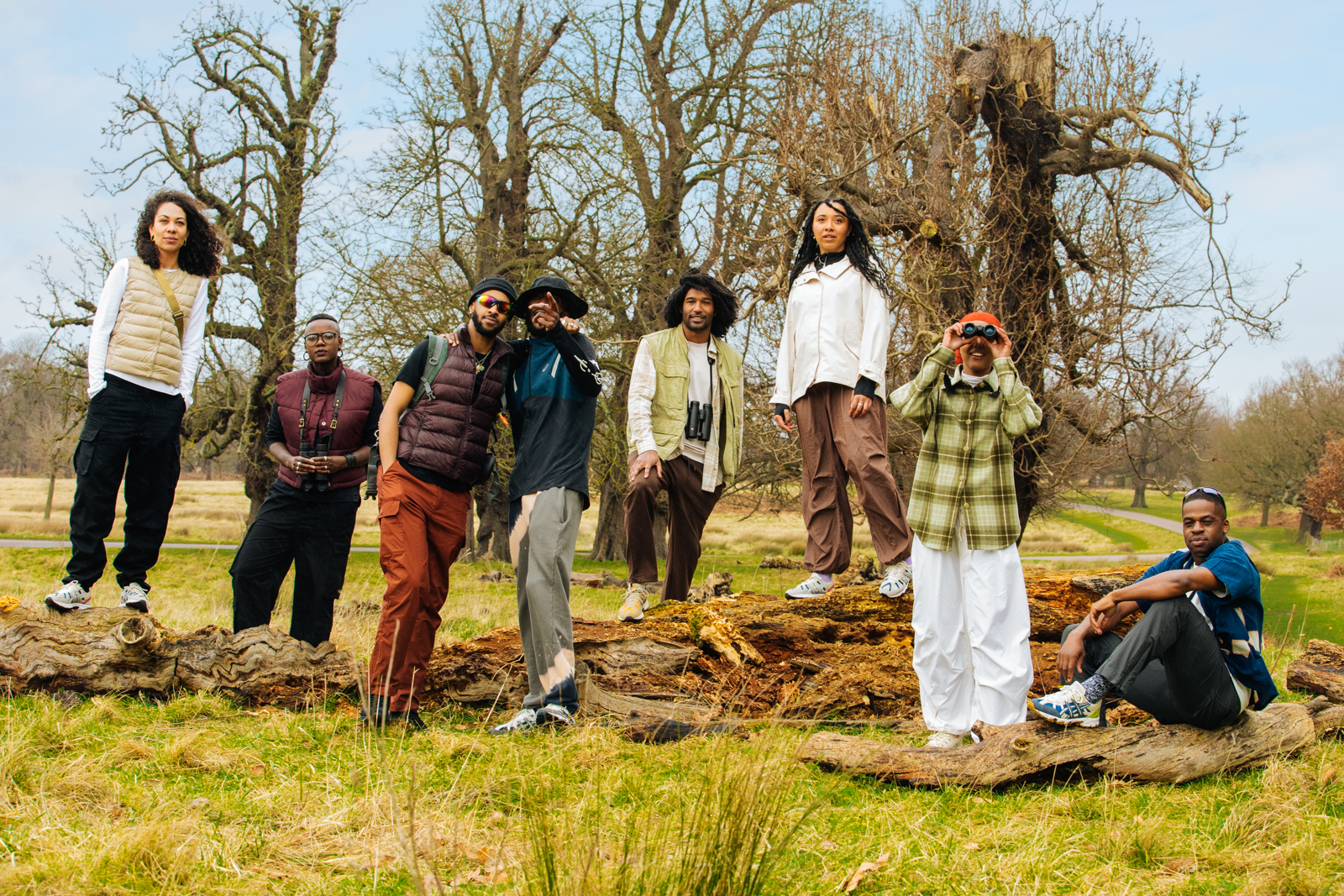 Group of seven adults in outdoor apparel standing and sitting on fallen tree trunk in park setting with bare trees in the background.