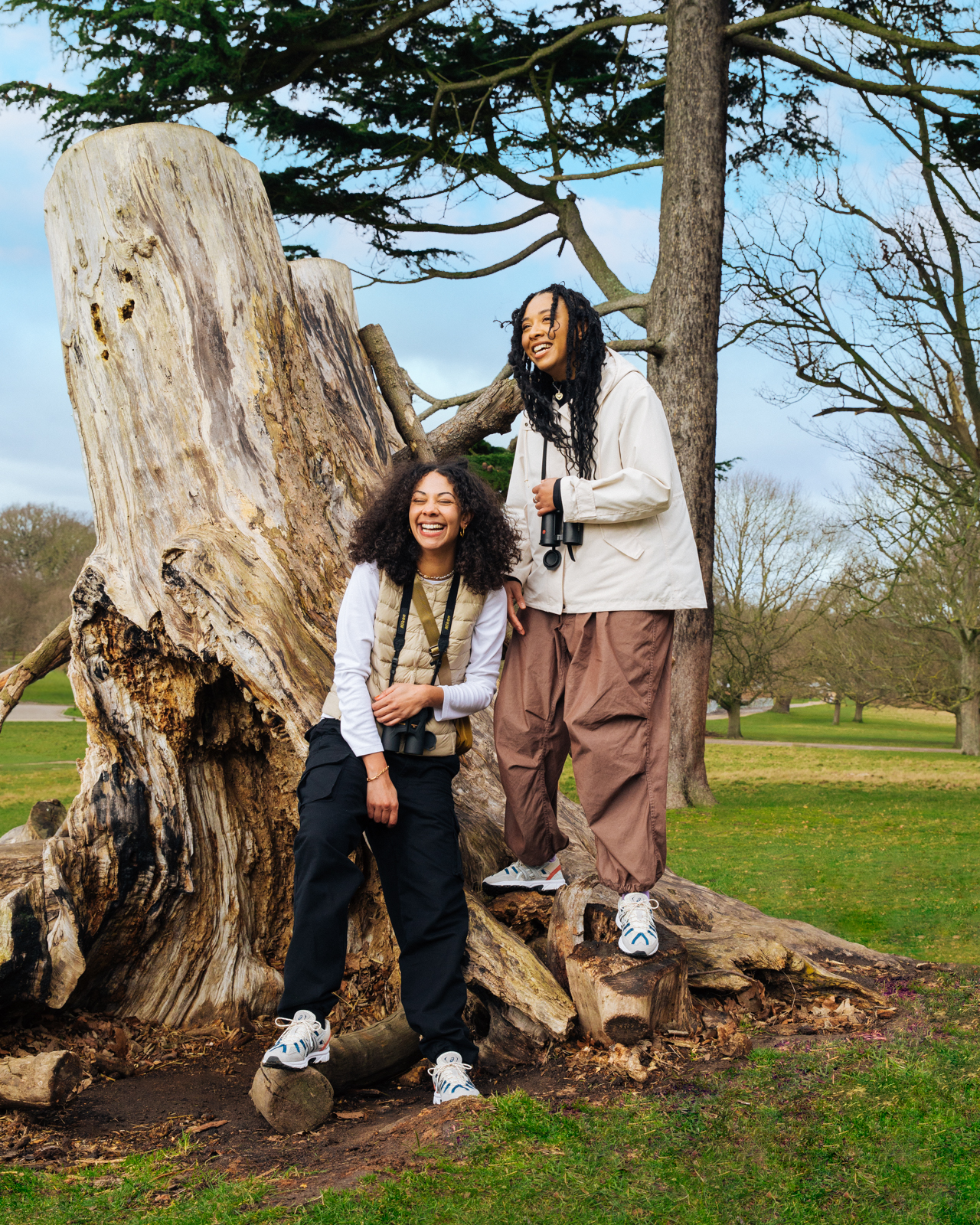 Two smiling women in outdoor apparel pose near a large tree trunk in a park setting on a sunny day, showcasing fashion and nature.