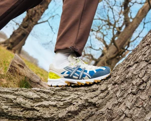 Close-up of ASICS running shoe on a tree trunk wearing brown trousers and white socks in a woodland setting
