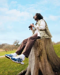 Man with curly hair seated on a tree stump holding binoculars, wearing a utility vest and blue sneakers in an outdoor landscape.