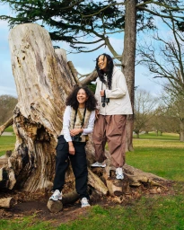 Two smiling women standing by a large weathered tree trunk in a park wearing utility apparel, vests, and running sneakers.