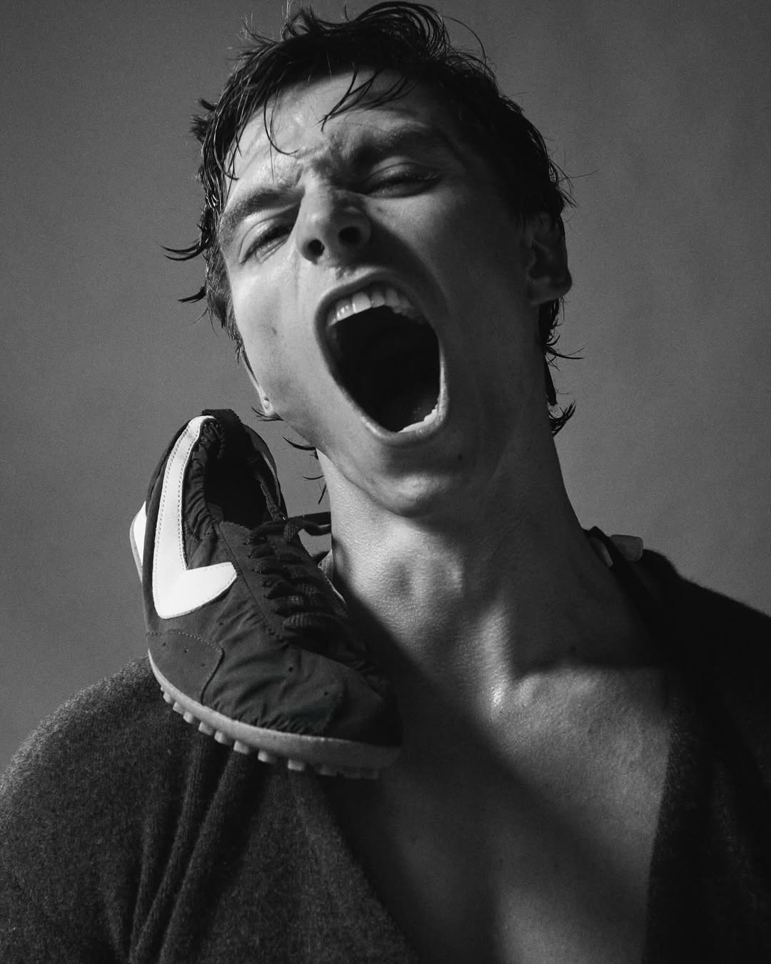 Monochrome portrait of a man with an open mouth, his sweaty hair disheveled, with a black and white sneaker resting on his shoulder.