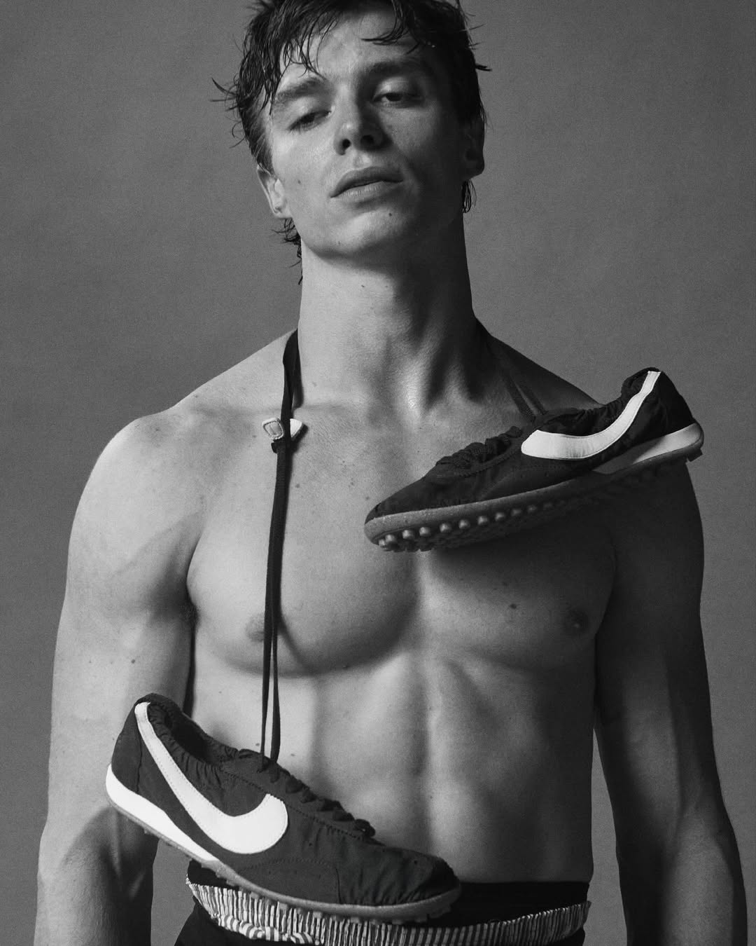 Black and white studio shot of a shirtless male model with black and white sneakers draped around his neck and waist against a grey backdrop.
