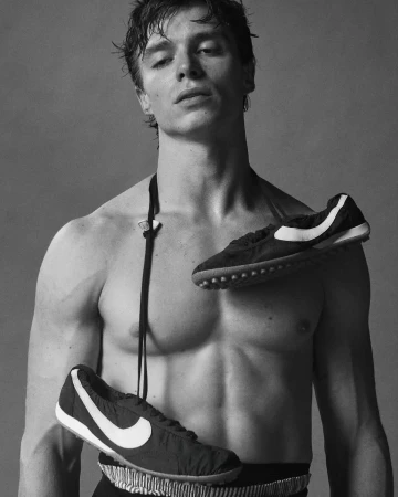Black and white studio shot of a shirtless male model with black and white sneakers draped around his neck and waist against a grey backdrop.