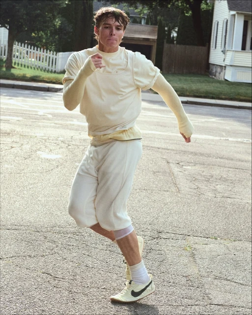 Young man wearing athletic clothing runs on an asphalt road. Residential houses and green foliage are visible in the background.