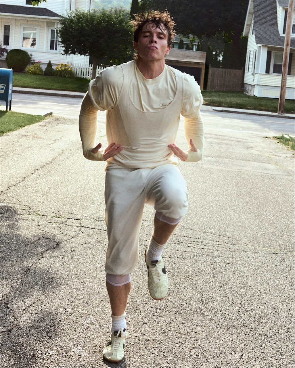 Athletic man in vintage athletic wear poses on a sunlit suburban street, wearing cleats and shorts, with houses and a mailbox in the background.