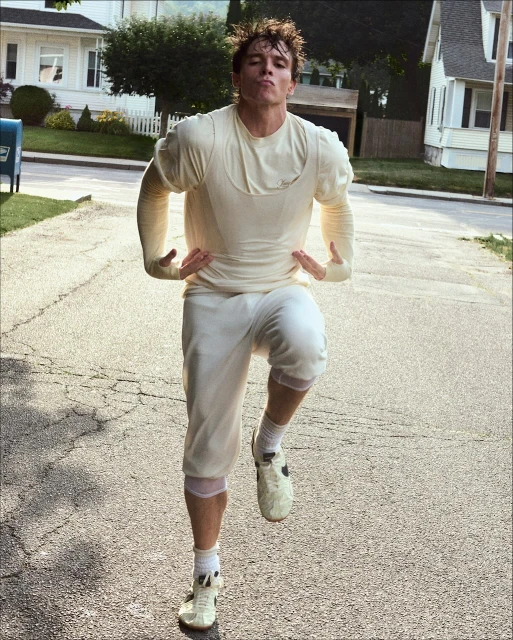 Athletic man in vintage athletic wear poses on a sunlit suburban street, wearing cleats and shorts, with houses and a mailbox in the background.