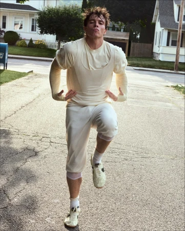 Athletic man in vintage athletic wear poses on a sunlit suburban street, wearing cleats and shorts, with houses and a mailbox in the background.