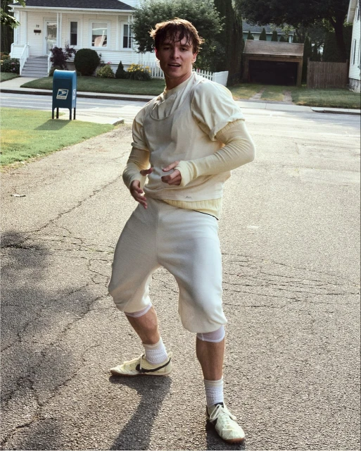 Young man in vintage football uniform stands on a suburban street with a mailbox and houses in the background, captured in natural light.
