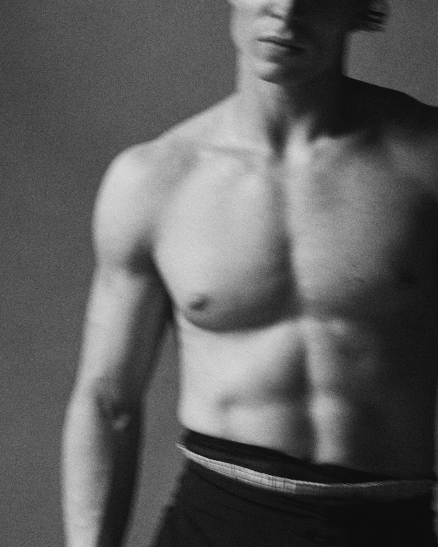 Monochrome studio shot of a muscular adult male torso with visible abs and chest, posing against a neutral gray backdrop.