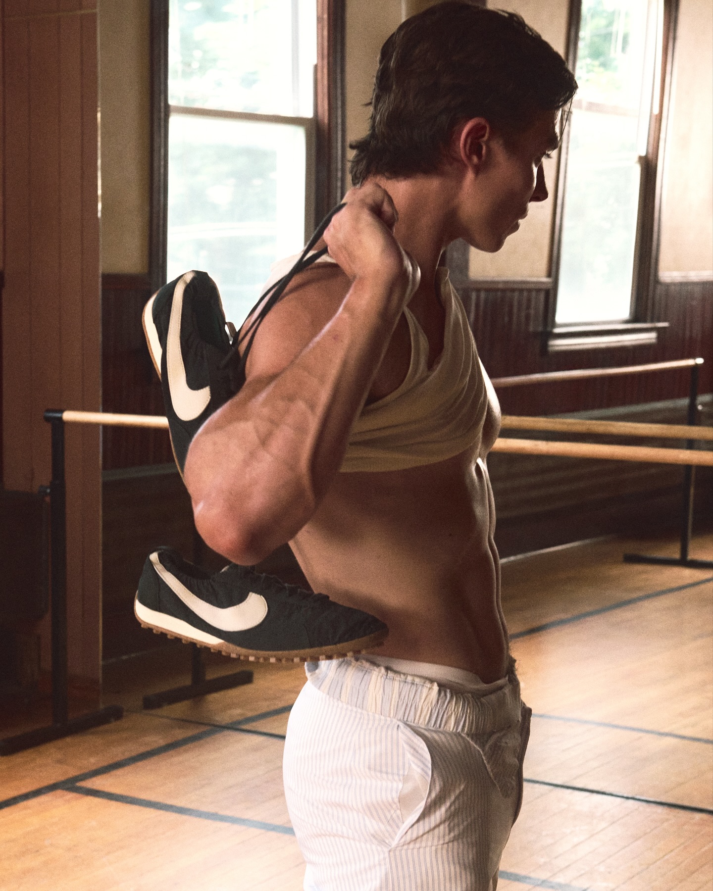 A man stands indoors in athletic gear, including a tank top and striped trousers, holding a pair of black Nike sneakers over his shoulder.