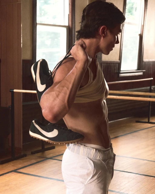 A man stands indoors in athletic gear, including a tank top and striped trousers, holding a pair of black Nike sneakers over his shoulder.