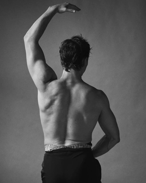 Back view of muscular man with arm raised. Black and white portrait of an adult male posing in dark pants against a neutral background.