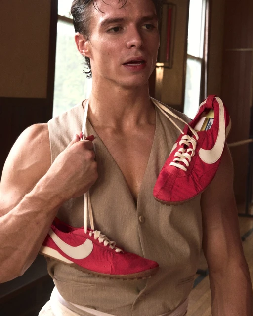A young man in a vest carries a pair of red and white Nike sneakers with laces around his neck, photographed indoors with soft lighting.