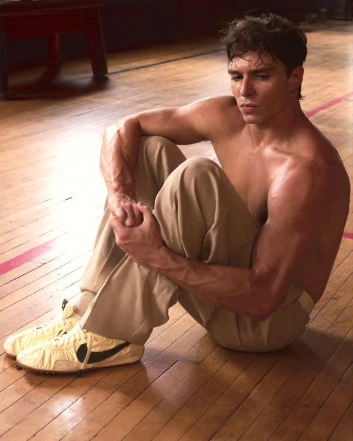 Muscular man sits shirtless on wooden floor with knees bent, wearing tan pants and vintage sneakers in indoor setting with natural light.